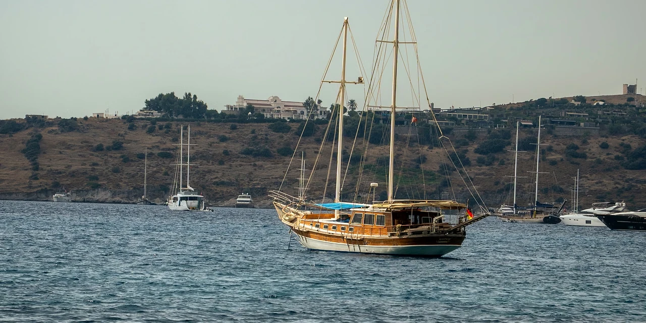 A wooden two-masted gulet at anchor off Bodrum, a dry hillside dotted with villas behind